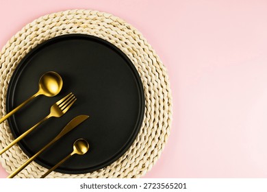 Top view of brown wicker table mat, black plate, gold cutlery on pink background. Straw round tray, flat lay, copy space. 
