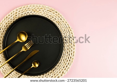 Top view of brown wicker table mat, black plate, gold cutlery on pink background. Straw round tray, flat lay, copy space. 