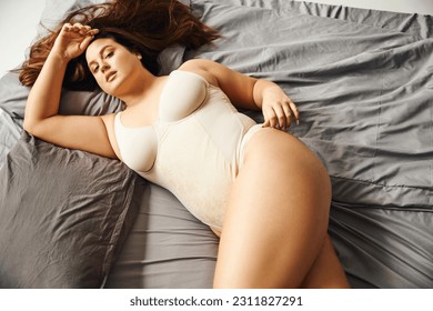 top view of brunette and curvy woman with natural makeup wearing beige bodysuit and lying on bed with grey bedding while looking at camera, body positive, figure type, modern apartment