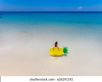 Top view of brunette woman standing on the beach with inflatable big pineapple in the transparent blue sea. Aerial view of lady relaxing on the floating mattress. 
