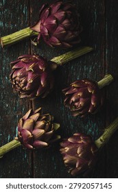 Top view of a bunch of five raw purple artichokes with stems arranged on a dark rustic wooden table with peeling teal paint. Organic Mediterranean food composition.