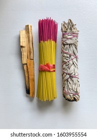 A top view of a bundle of yellow and redish purple incense tied with a pink ribbon with sticks of palo santo lying in vertical rows to the left and a bundle of dried sage to the right on