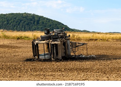 Top View Of A Burnt Out Hay Baler Lying In A Field Against A Green Forest With The Field Around It Having Been Plowed To Prevent Spread Of Fire To The Dry Grass In Background