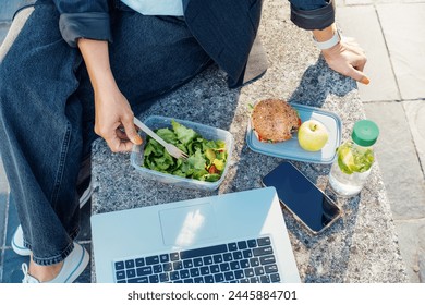 Top view business woman having lunch break, eating fast and healthy in front of laptop outdoors. Balanced diet lunchbox with fresh salad, sandwich, apple, water. Healthy eating habits and well-being. - Powered by Shutterstock