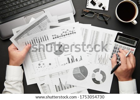Top view of businessman's hands working with financial reports. Modern black office desk with laptop, notebook, pencil and a lot of things. Flat lay table layout.