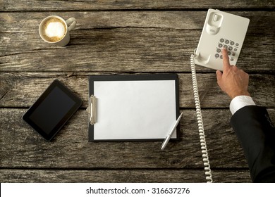 Top View Of Businessperson Making A Call With His Office Phone With Digital Tablet And Blank Piece Of Paper On A Rustic Wooden Desk. Concept Of Telemarketing, Feedback And Customer Support.