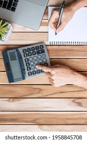 Top View Businesswoman Hand Writing And Calculating With Calculator, Pen, Computer Laptop And Plant Pot On Wood Office Desk Table Background. Workspace Or Home Office With Copy Space For Text Concept