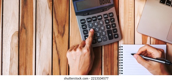 Top View Businesswoman Hand Writing And Calculating With Calculator, Pen, Computer Laptop And Plant Pot On Wood Office Desk Table Background. Workspace Or Home Office With Copy Space For Text Concept