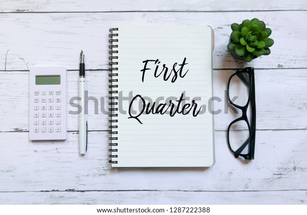 Top view of calculator,glasses,pen,plant and notebook written with First Quarter on wooden background.Business and finance concept.