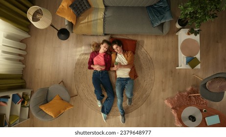 Top view capturing a young couple laying on the floor next to each other, looking at the screen of the smartphone as if they were watching pictures.