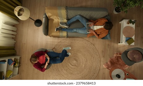 Top view capturing a young man laying on the couch with a laptop screen while his girlfriend is sitting on a chair next to him with a smartphone.