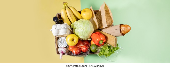 Top View Of Cardboard Box With Food Products On Color Green-yellow Background. Safe Delivery. Contactless Home Food Delivery. Stock For Rainy Day. Food Donation.  Banner With Copy Space.