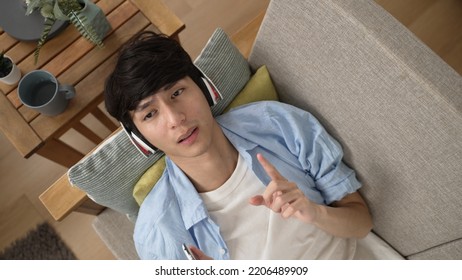 Top View Carefree Asian Chinese Male Lying On Sofa Is Singing And Moving Finger As A Conductor While Enjoying Music From Headphones In The Living Room At Home.