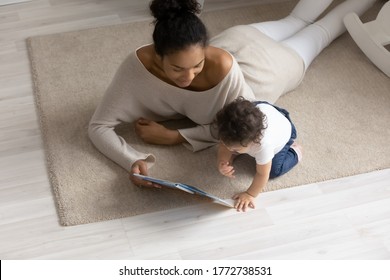 Top View Caring African American Mother Reading Book To Toddler Daughter, Lying On Warm Floor With Underfloor Heating, Loving Mum And Cute Little Girl Playing, Enjoying Leisure Time Together