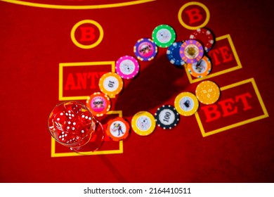 Top View Of Casino Chips And A Glass Filled With Red Dice. A Set Of Dice And Playing Chips For Craps, Poker, Roulette On A Red Gaming Table In A Casino. Close Up. Background Of Gambling.