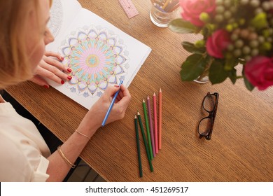 Top View Of Caucasian Female Drawing In Adult Coloring Book. Glasses And Color Pencils On Wooden Table At Home.