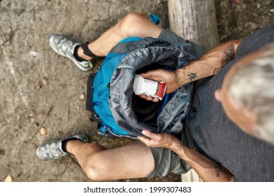 Top View Of Caucasian Man Holding Snack Pack Above Open Backpack While Resting On Log In Sunny Nature, Selective Focus. Hiking, Travel Outdoor, Recreation And Active Adventure Concept