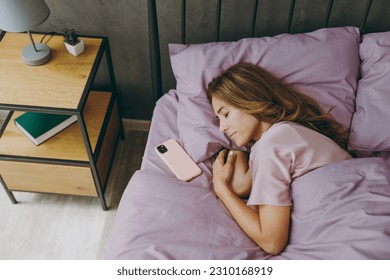 Top view caucasian young woman wear purple t-shirt pajama lying in bed sleep near mobile cell phone rest relax spend time in bedroom lounge home in own room hotel wake up dream. Real estate concept - Powered by Shutterstock