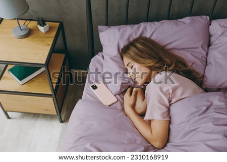 Top view caucasian young woman wear purple t-shirt pajama lying in bed sleep near mobile cell phone rest relax spend time in bedroom lounge home in own room hotel wake up dream. Real estate concept