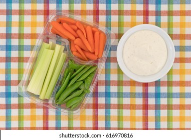 Top View Of Celery, Carrots And Sugar Snap Peas In A Plastic Container With A Bowl Of Ranch Dressing To The Side Atop A Colorful Cloth Patterned Place Mat.