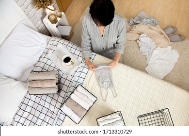 Top view of cheerful beautiful young brunette woman tidying up things in her wardrobe while sitting on floor near bed in stylish bedroom with cup of coffee. Spring tidy concept