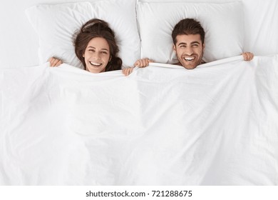 Top view of cheerful excited Caucasian married couple lying in bed hiding bodies under white blanket, looking at camera and smiling happily, waking up in hotel room after sleepless wedding night