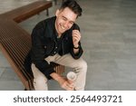 Top view of cheerful man sitting on city bench, enjoying coffee break in vibrant urban setting, laughing while savoring moment and embracing simple pleasures of life. Concept of urban lifestyle.