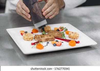 Top View Of The Chef's Hands Carefully Pouring Sauce On The Finished And Beautifully Sorted Dish Of Fresh Grilled Meat With Rice And Slices Of Fruits And Greens On A White Plate And Table