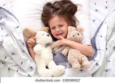 Top view of child crying in bed, sad kid on pillow in bedroom, liitle girl lying on linens with dandelion, upset toddler in pajama lay swith her teddy bears in bed in morning. Childhood concept.