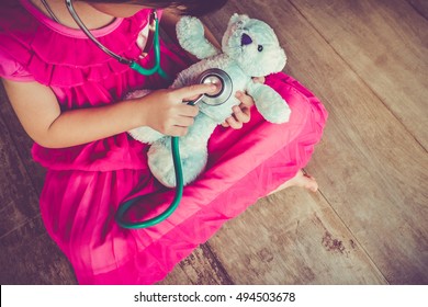 Top View Of Child Playing Doctor Or Nurse With Plush Toy Bear With Bright Sunlight At Home. Happy Girl Listens A Stethoscope To Toy. Playful Girl Role Playing. Vintage Tone Effect.