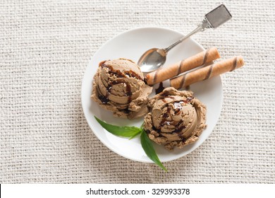 Top View Chocolate Ice Cream With Wafer And Spoon On Plate, Table Cloth Background.