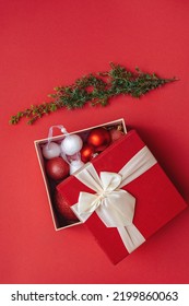 Top View Of Christmas Decorations In Open Gift Box And Green Christmas Tree Branch On Plain Background. Red Packaging Lid With Lush Bow. Merry Christmas And Happy New Year. Copy Space. Vertical Photo