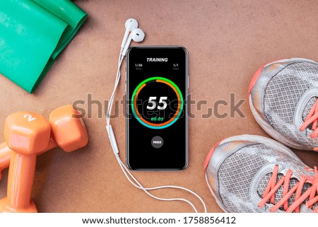 a top view of a chromometer app on a black phone with headphones above a blue mask, a test tube blood, dumbbells. measure tape, shoes for workout at home