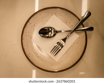 Top view of a clean dining setup with a spoon and fork placed on a napkin over a speckled ceramic plate. Perfect for restaurant, culinary, or hospitality-themed visuals.