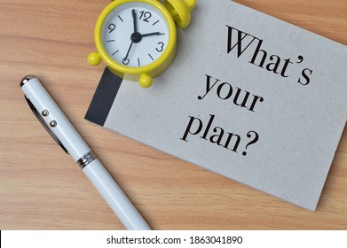 Top view of clock, pen and notebook over wooden background written with question WHAT'S YOUR PLAN?. Business and education concept.