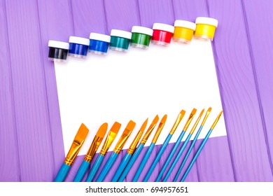 Top view of close cans of colored paint with brushes and sheet of paper on the purple wooden background