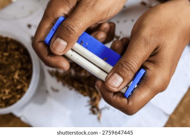 Top view close up of hands manually rolling a cigarette using a small blue rolling machine, with loose dried tobacco spread on a white surface next to a container filled with tobacco.