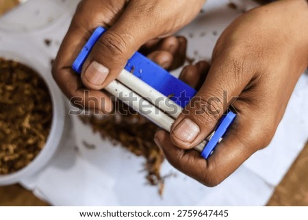 Top view close up of hands manually rolling a cigarette using a small blue rolling machine, with loose dried tobacco spread on a white surface next to a container filled with tobacco.