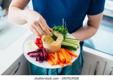 Top view close up man's hand dipping cucumber stick in hummus on the kitchen. Hummus served with raw vegetables on the plate. Healthy food lunch. Vegetarian and vegan food diet. Soft selective focus