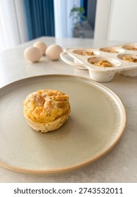 Top view close up of a single delicious homemade egg muffin with golden crust served on a ceramic plate. Healthy high protein breakfast set in a bright minimalist kitchen with natural morning sunlight and aesthetic hard shadows. Background features blurred whole eggs and a silicone baking dish, creating a cozy home cooking atmosphere. Ideal for ketogenic diet, weight loss, and anti-aging lifestyle content. Savory crustless quiche appetizer, nutritious meal prep, and clean eating recipe concept on a light stone texture table with copy space