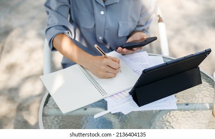 Top View And Close Up, Hand Of Asian Woman Writing Her Plan On Notebook And Using Mobile Phone With Tablet Computer Working Online With Paperwork Of Investment On Glass Table