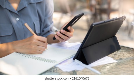 Top View And Close Up, Hand Of Asian Woman Writing Her Plan On Notebook And Using Mobile Phone With Tablet Computer Working Online With Paperwork Of Investment On Glass Table