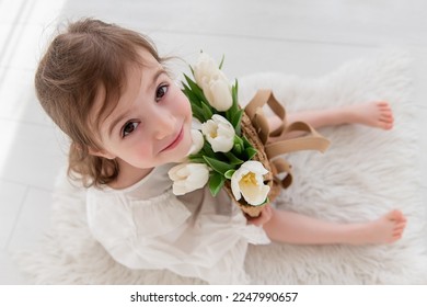 Top view close-up portrait of little girl in a white dress, nightgown. Toddler embraces a bouquet of fresh, delicate white tulips. Gift for the holiday, the concept of purity, spring time. Copy space - Shutterstock ID 2247990657