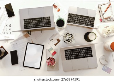 Top view of a cluttered workspace with three laptops, a tablet, smartphones, coffee, and office supplies on a white desk.