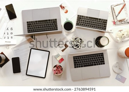 Top view of a cluttered workspace with three laptops, a tablet, smartphones, coffee, and office supplies on a white desk.