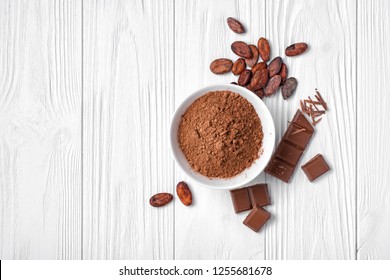 Top view of cocoa powder with broken chocolate bar and cocoa beans for confectionery on white wooden background with copy space