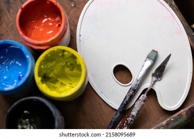 Top View Of Colored Acrylic Paint Pots, Plastic Palette, Brush And Palette Knife Used For Painting In The Art Studio. Space For Copying. Concept Of Artist And Hobby