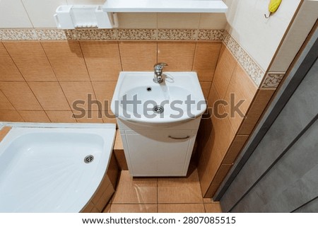 Top view of compact bathroom corner showing white ceramic sink on slim storage cabinet beside bathtub edge, warm brown wall and floor tiles, floral border, chrome fixtures, clean modern interior