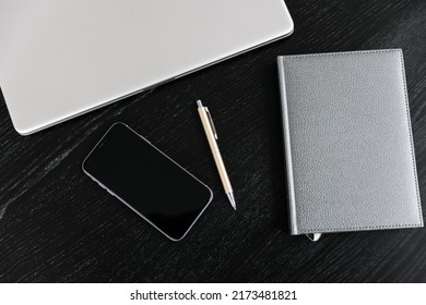 Top view of the composition against a background of ebony. Closed diary with gray leather cover, gold pen, closed silver notepad, phone. Desk in the office.  Free space for text. Space for copying.