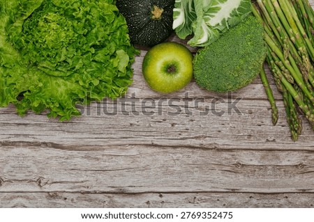 Top view composition featuring vibrant assortment of green produce: lettuce, broccoli, asparagus, round zucchini, and green apple. The vegetables are arranged along the top edge, creating a natural border on a rustic grey-toned wooden plank surface. The bottom half of the image provides large, clean area for copy space.